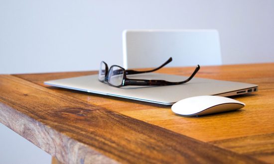 Laptop, Brille und Maus auf einem Holztisch, im Hintergrund ein Stuhl.